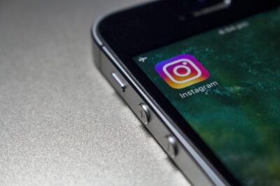 Das Bild zeigt ein Smartphone, auf dessen Bildschirm die Instagram-App geöffnet ist. Das Instagram-Symbol ist prominent sichtbar, mit einem bunten Kameralogo auf einem grünlichen Hintergrund. Das Gerät liegt auf einer strukturierten, grauen Oberfläche. Dieses Bild könnte auf der Website von 420MKT, einer Cannabis-Marketing-Agentur, verwendet werden, um den Einsatz von Social Media für Cannabis- oder CBD-Marketing zu illustrieren. Schlagwörter wie Social Media, Instagram und Marketing sind hier relevant.